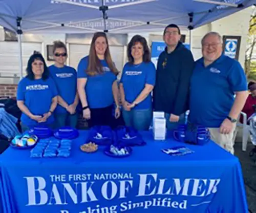 Group of adults all in blue shirts posing for photo behind blue table with Bank of Elmer banner