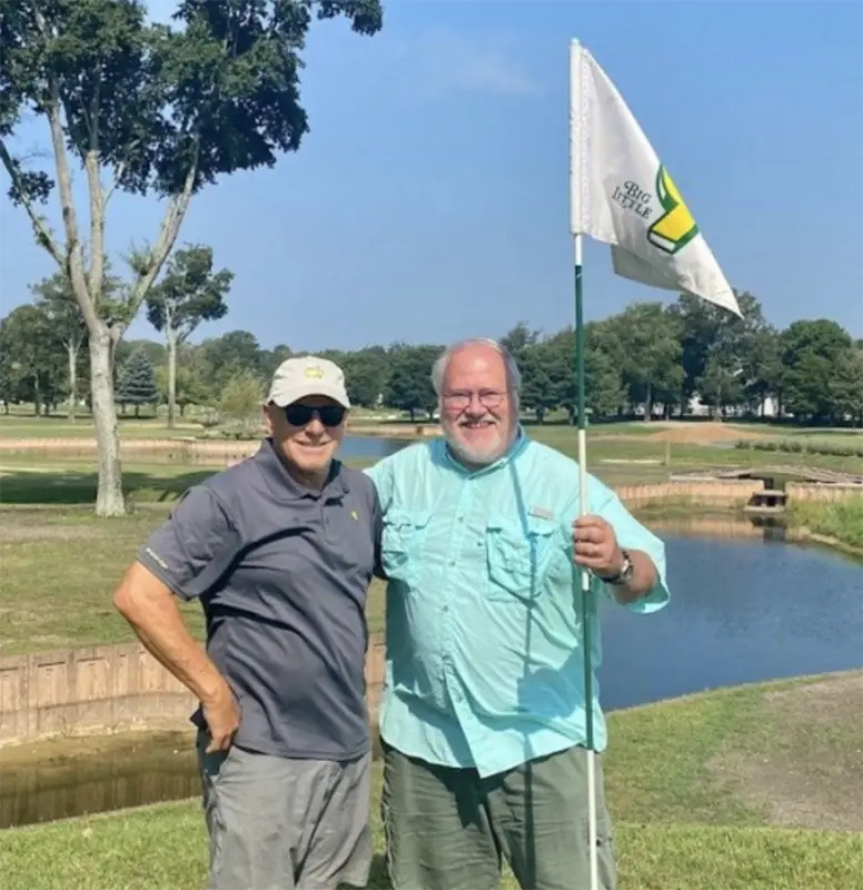 Two large men standing, holding a golf flag